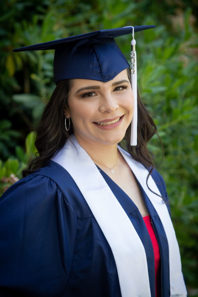 woman with long black hair wearing blue cap and gown and white stole smiling